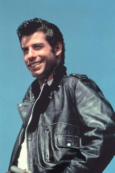 Promotional still of a man in a black leather motorcycle jacket smiling under a blue sky